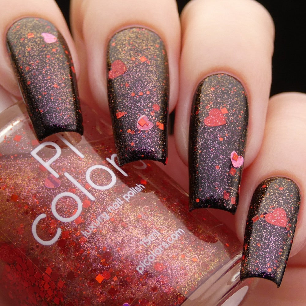 A close-up shot captures a hand with long nails adorned in a mesmerizing black glitter polish, featuring red holographic hearts. The hand holds a bottle of PI Colors' Strawberry Heart.208 Red Nail Polish Topper, highlighting the captivating red glitter inside. The softly blurred background beautifully complements the striking manicure.