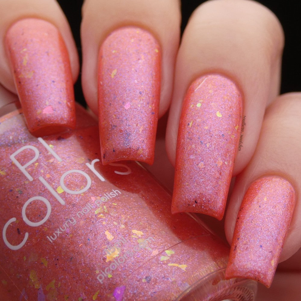 A close-up of four fingers reveals nails painted in Lala Ngoxolo Kumkani Coral Nail Polish by PI Colors, with shimmering pink and iridescent flake accents. Partially visible, the bottle exudes a glamorous appeal as it is held by the fingers.