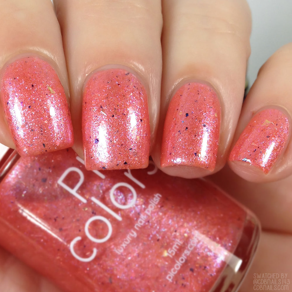 Close-up of a hand with short nails adorned in a shimmering peachy coral nail polish. The custom handmade "Lala Ngoxolo Kumkani Coral Nail Polish" by PI Colors features iridescent rainbow flakes, adding a colorful touch. The bottle is visible next to the painted nails, showcasing its unique charm.