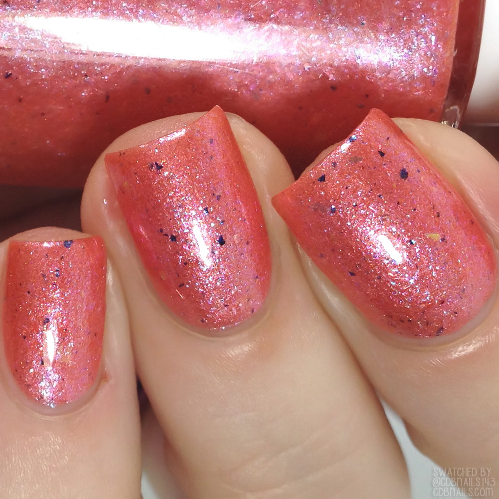 Close-up of nails painted with Lala Ngoxolo Kumkani Coral Nail Polish by PI Colors, showcasing a pinkish-red glittery hue. The shimmering finish and small dark speckles create a textured appearance, while iridescent rainbow flakes add depth. In the background, a matching bottle of this unique nail polish is visible.