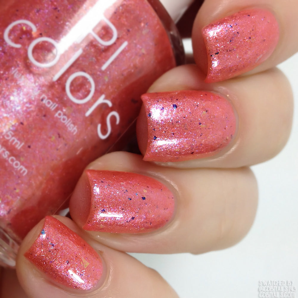 A close-up of a hand features nails painted with Lala Ngoxolo Kumkani Coral Nail Polish, adorned with iridescent rainbow flakes. The other hand holds a bottle labeled "PI Colors," highlighting a glossy finish with delicate blue and silver flecks.