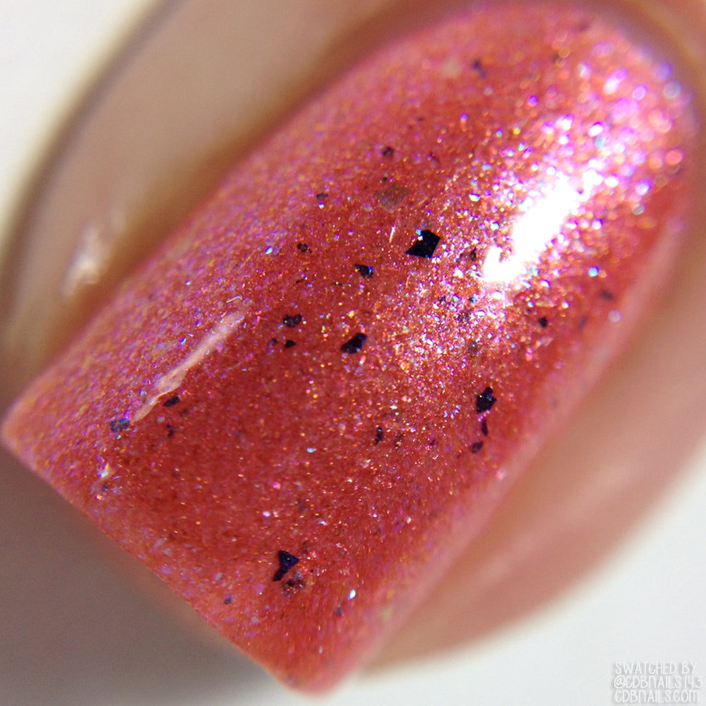Close-up of a fingernail adorned with PI Colors' Lala Ngoxolo Kumkani Coral Nail Polish, a custom handmade formulation in shimmery pink that includes tiny black specks and iridescent glitter. The glitter reflects light to create a shiny, sparkling effect on a well-manicured nail.