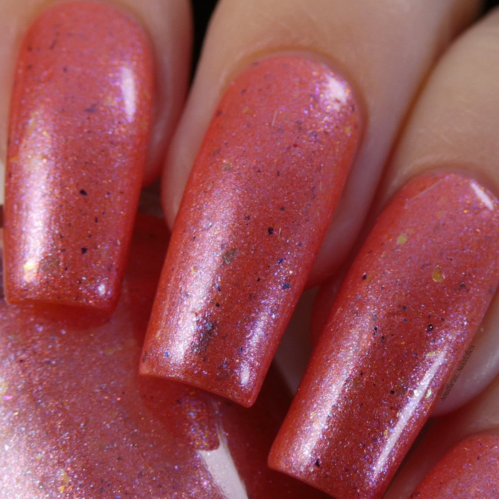 A close-up of a hand with long fingernails painted in PI Colors' Lala Ngoxolo Kumkani Coral Nail Polish shows off a stunning iridescent pink that glistens with tiny specks of glitter and iridescent rainbow flakes, giving the nails a vibrant, sparkling appearance.