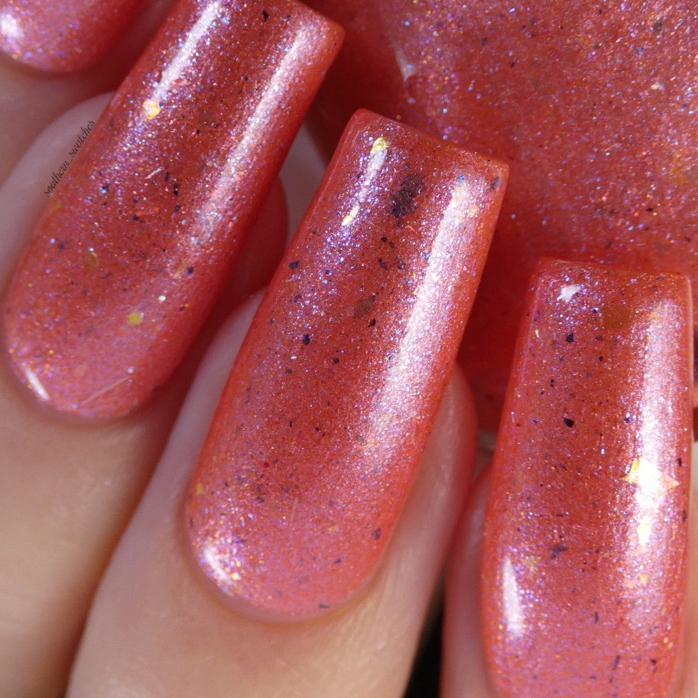A close-up of fingers with long nails painted in PI Colors' Lala Ngoxolo Kumkani Coral Nail Polish, a custom handcrafted shimmery coral featuring black speckles and touches of gold. Iridescent rainbow flakes sparkle in the light, set against a backdrop that mirrors the glittery coral surface.