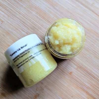Two jars of Lemony Sugar Lip Scrub by PI Colors rest on a wooden surface. One jar showcases its list of organic ingredients, highlighting the nourishing rosehip oil, while the other is open, revealing the textured yellow scrub within.