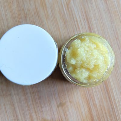 A small open jar of PI Colors' Lemony Sugar Lip Scrub, featuring a white lid, rests on a wooden surface. Made with organic ingredients, the scrub has a grainy texture and contains nourishing rosehip oil.