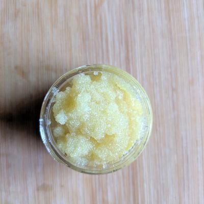 A petite jar bearing the "Lemony Sugar Lip Scrub" label from PI Colors rests on a wooden surface, brimming with a granular, yellow substance. Its coarse and grainy texture offers exfoliation using organic ingredients to achieve the softest lips.