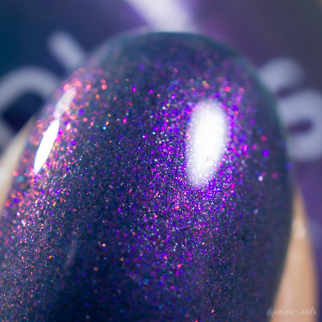 A close-up of a fingernail adorned with PI Colors' Night Terrors.000 Violet Nail Polish reveals a glossy, color-shifting shimmer. The hue appears purple, accentuated by tiny pink and blue glitter particles that create a sparkling effect. This 10-free polish beautifully reflects light, producing a shiny highlight.