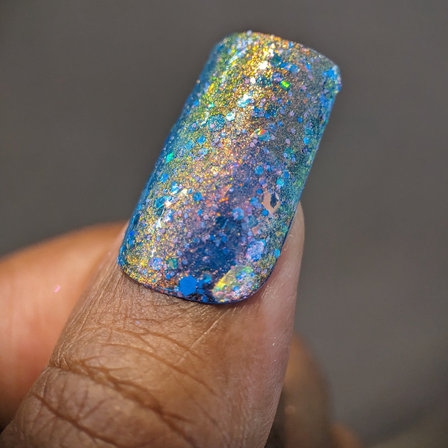 A close-up of a finger adorned with PI Colors' Rainbow Quartz.000 nail polish reveals a dazzling multichrome effect. The vibrant sparkles include a stunning blend of blue, gold, green, and pink hues, creating an iridescent appearance. Metallic blue holo glitter enhances the depth against the soft-focus gray background.