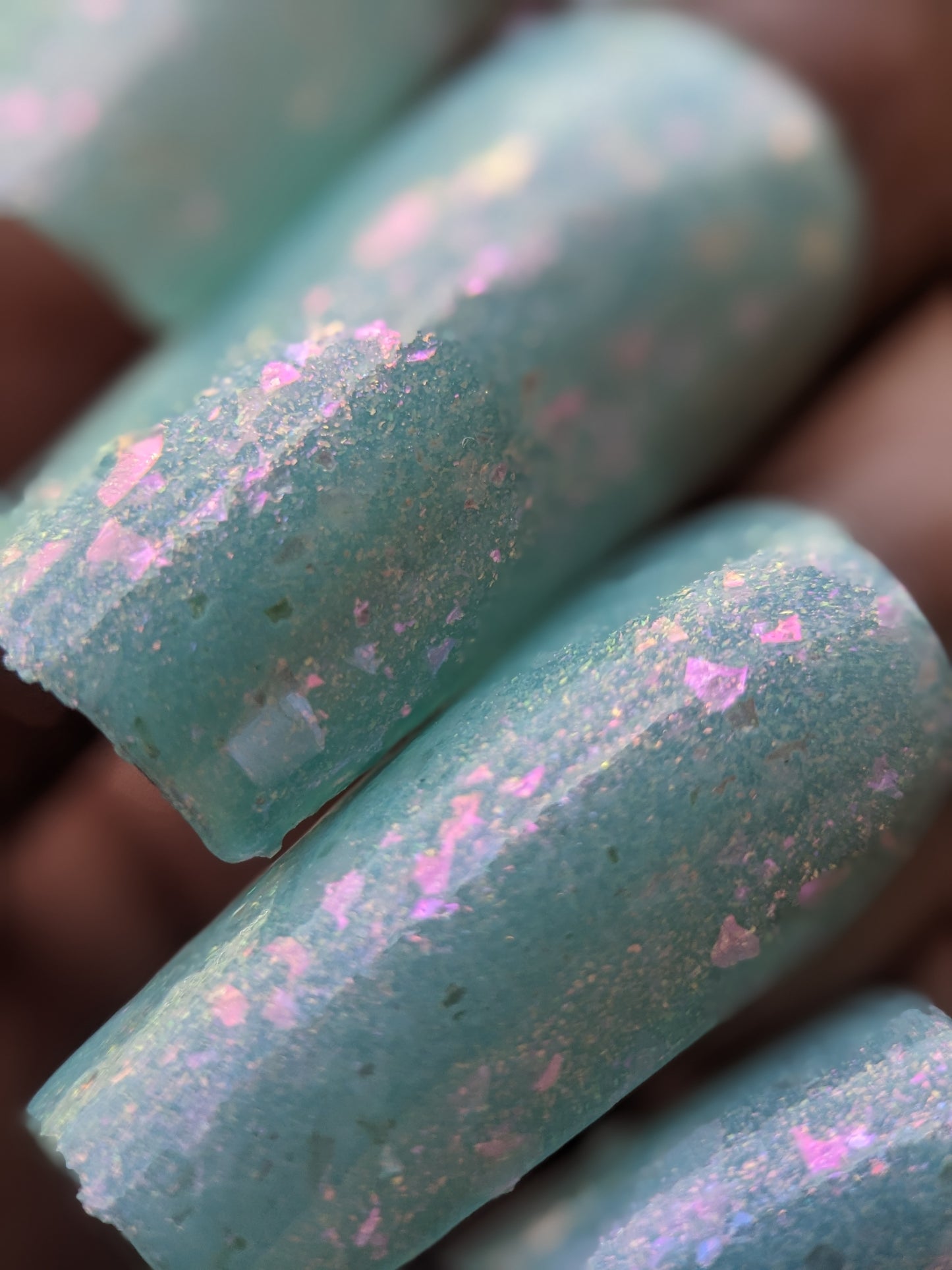Close-up of the Star of Siam.000 nail polish by PI Colors, featuring pastel teal with color-shifting rainbow flakies that create a textured and shimmering effect. The neatly manicured nails reflect pink and purple hues in the light due to the glitter.