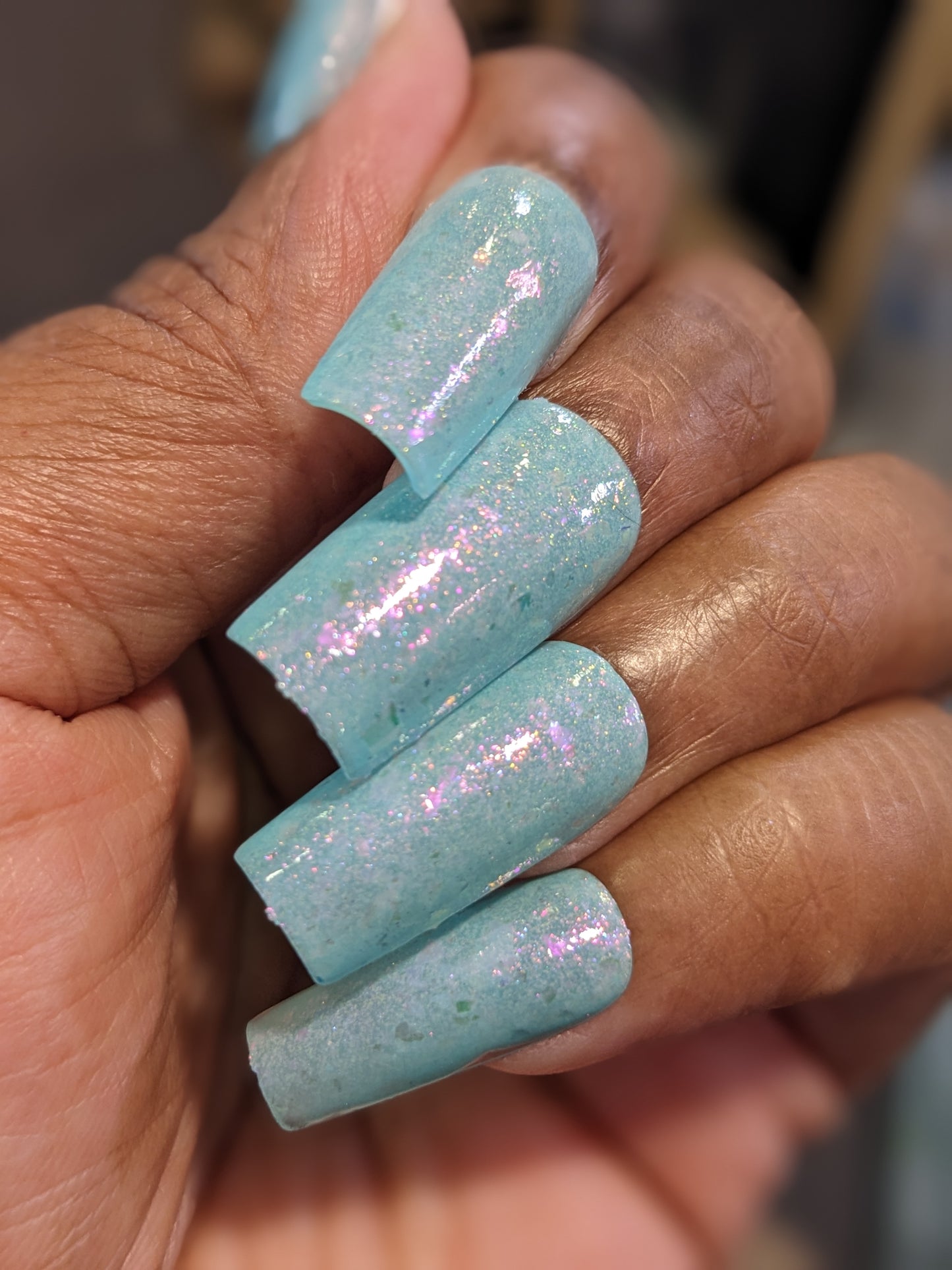 A hand showcases long, glossy nails painted in the Star of Siam.000 shade by PI Colors, featuring a shimmering opalescent finish with colorshifting rainbow flakies. The well-manicured nails have a slightly curved shape and highlight the beauty of this 10-free nail polish.