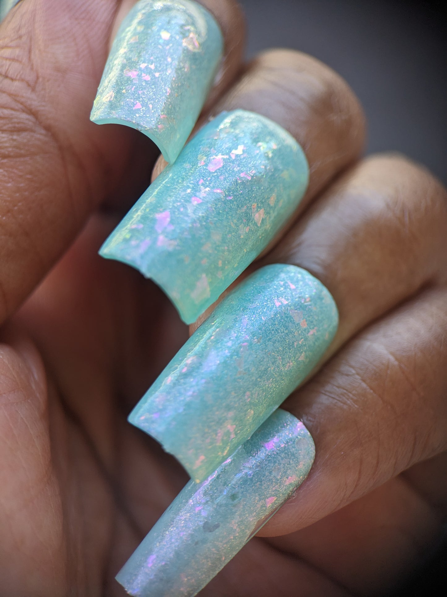 A close-up of a hand with long nails adorned with PI Colors' "Star of Siam.000" nail polish, showcasing a pastel teal hue with a shimmering, iridescent finish enriched with pink and white flecks. The glossy texture is further enhanced by colorshifting rainbow flakies, providing a mesmerizing, textured look. This 10-free polish from PI Colors truly captivates.