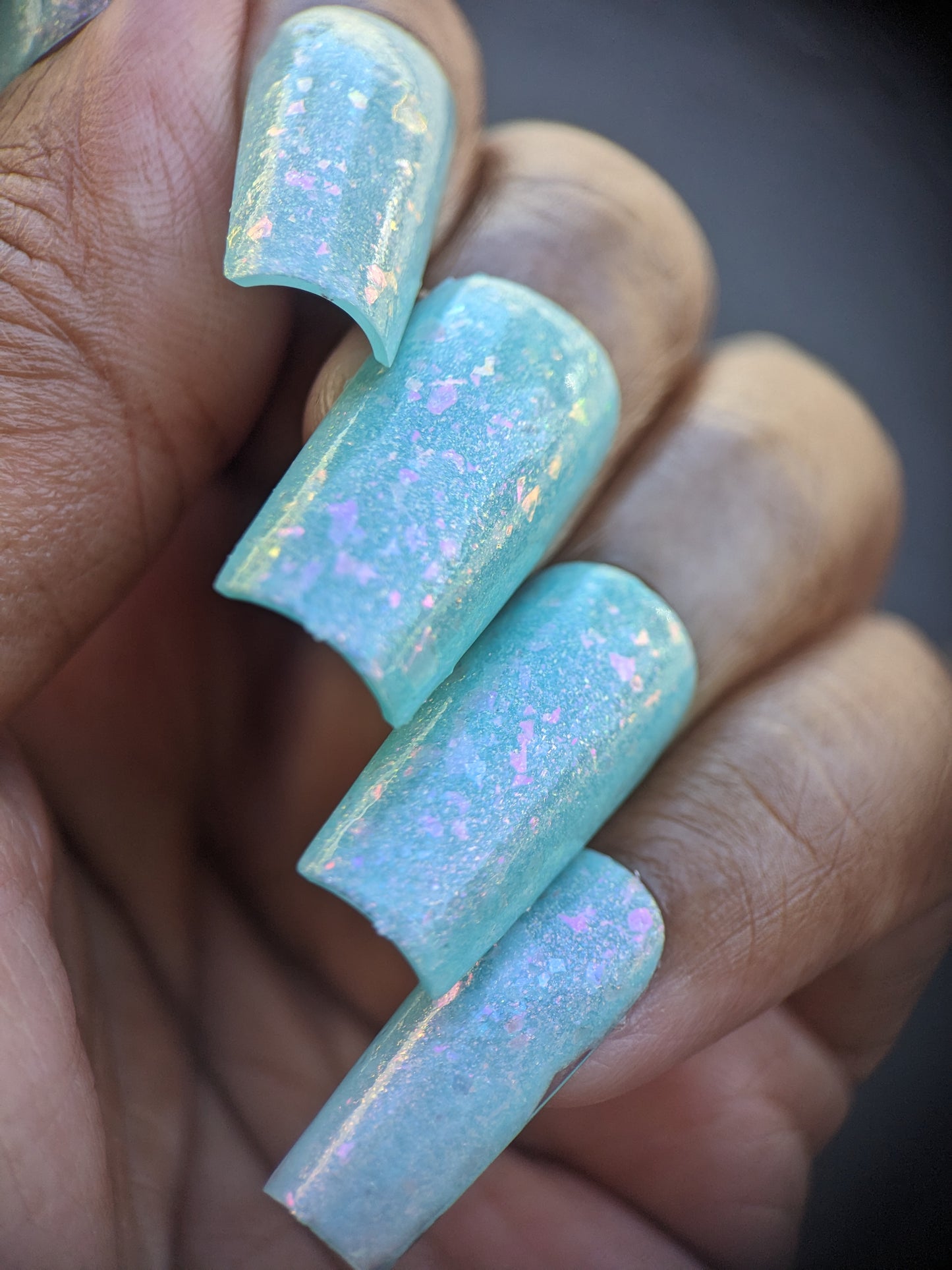 A close-up of a hand displays long nails painted in the pastel teal shade of PI Colors' Star of Siam.000 10-free nail polish. The impeccably manicured nails are embellished with iridescent, color-shifting rainbow flakies, providing a smooth, glossy finish that reflects different hues when viewed in varying light conditions.