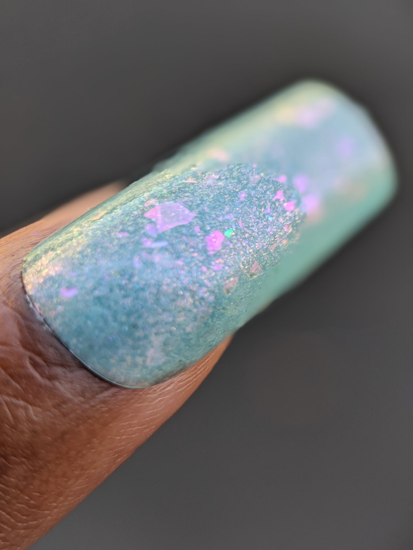 A close-up of a fingernail showcases the enchanting Star of Siam.000 from PI Colors, featuring a pastel teal shade enhanced with color-shifting rainbow flakes. The iridescent finish shimmers with pink and purple flecks, creating a multi-dimensional appearance. As a 10-free nail polish, it offers a vibrant and captivating look.