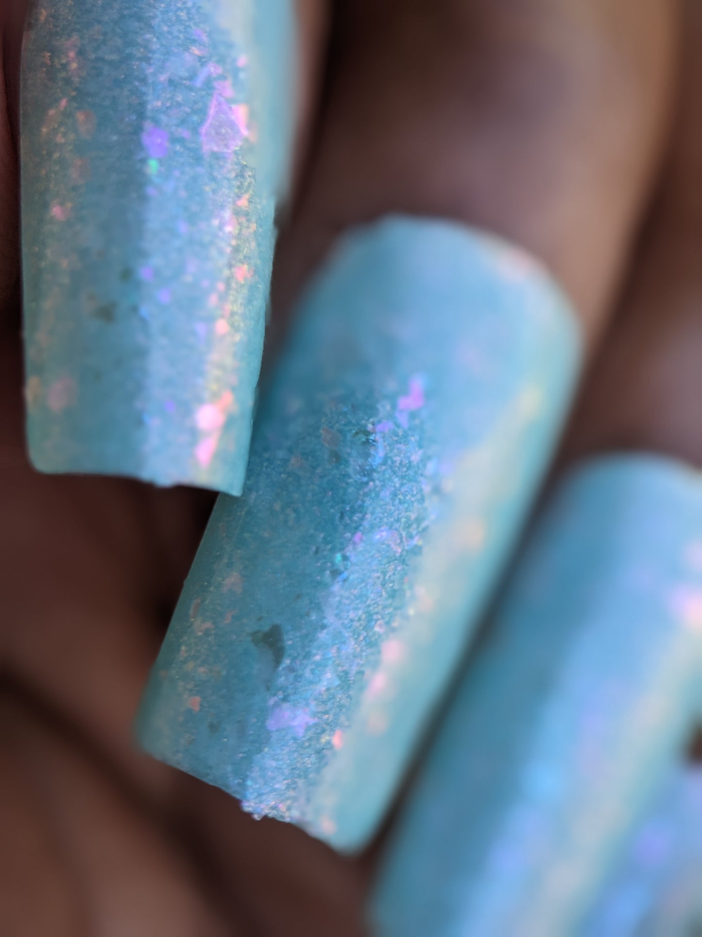Close-up of a hand with long, rectangular-shaped nails adorned in Star of Siam.000 by PI Colors—an exquisite 10-free nail polish boasting a textured turquoise shade with subtle pink flecks and dazzling color-shifting rainbow flakies. The lighting emphasizes the intricate details and shimmering finish beautifully.