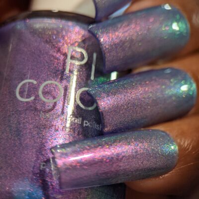 A close-up of a person's hand shows off long nails adorned with PI Colors' "Dusk Rainbow.321" purple nail polish, featuring a deep hue with mesmerizing cyan and green aurora shimmer. This handmade polish captures enchanting shades of pink, blue, and green. The partially visible bottle seamlessly complements the captivating nail color.