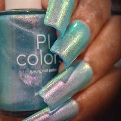 A close-up of a hand with long, iridescent aqua nails holding a matching bottle of PI Colors' Ocean Whitecap.309 Blue Nail Polish. This handmade polish features a shiny, reflective finish and exquisite color-changing effects that transition between hues of blue and purple.