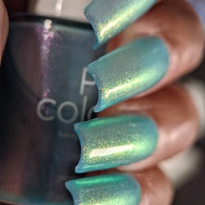 A close-up of a hand with long, shiny, iridescent turquoise nails showcases a bottle of PI Colors' Ocean Whitecap.309 Blue Nail Polish. This captivating nail polish features a mesmerizing blue-green hue that shifts to reveal hints of green and purple in the light.