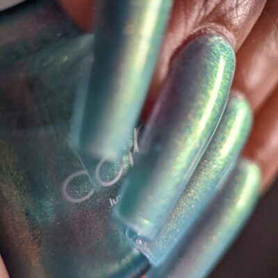 A close-up captures a hand with long, shimmering turquoise nails holding a bottle of PI Colors Ocean Whitecap.309 Blue Nail Polish. The nails exhibit an iridescent sheen, reflecting stunning shades of blue and green with mesmerizing color-changing effects.