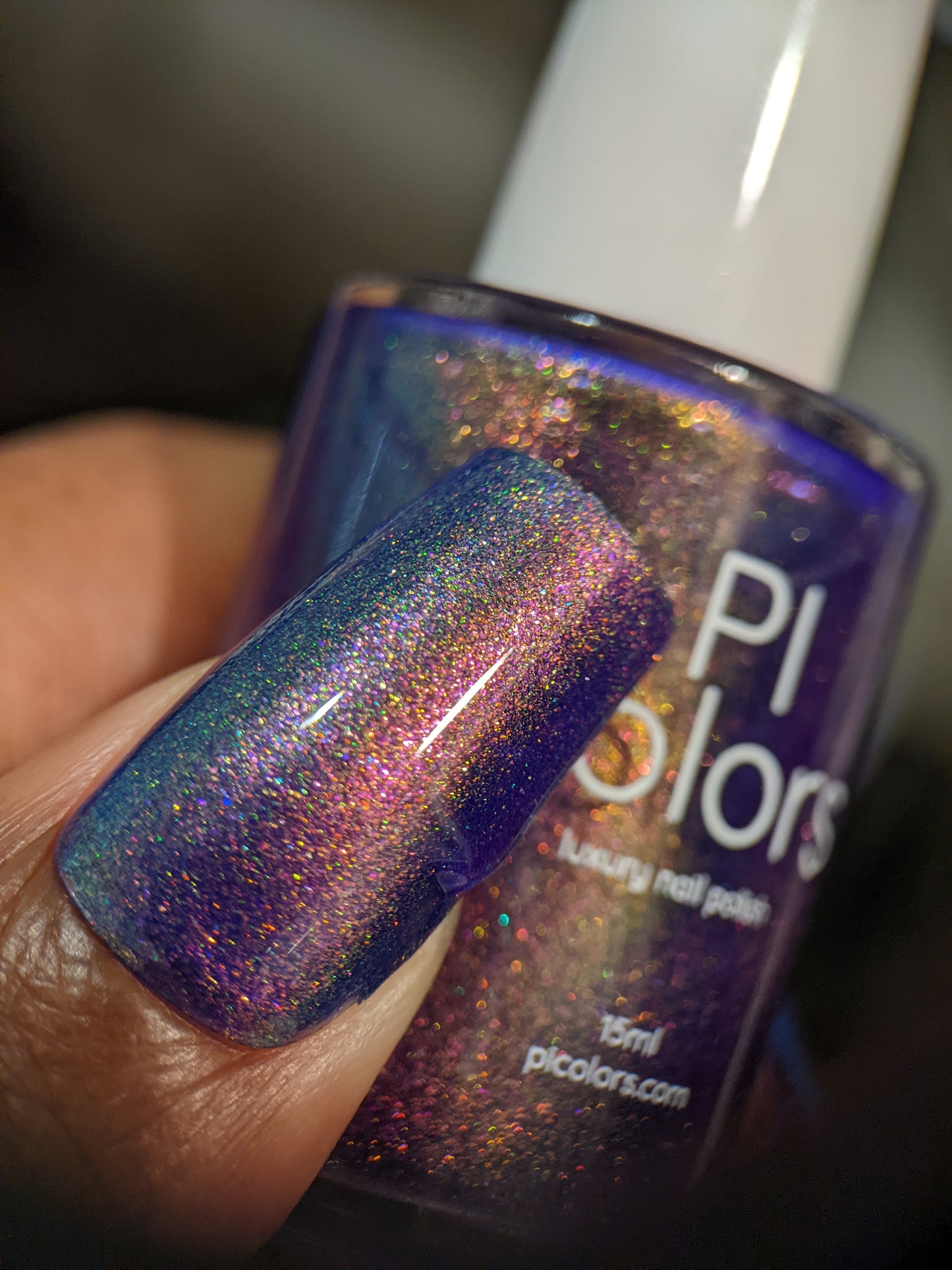 A close-up of a fingernail painted with multi-chrome holographic nail polish reveals a color transition from blue to violet purple. The fingernail holds a bottle of PI Colors' 90s Kind of World Purple Nail Polish, which features a similar shimmering hue enhanced with aurora pigment.