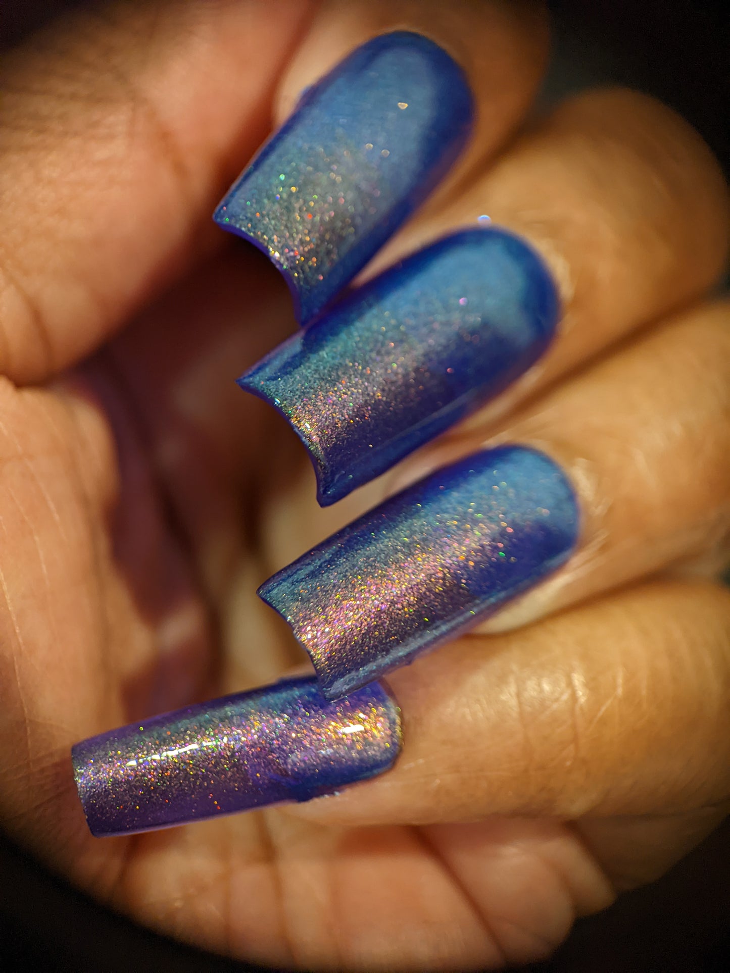 A close-up view highlights a hand adorned with long, shimmering nails painted using PI Colors' "90s Kind of World Purple Nail Polish." The vibrant mixture of blue and violet purple polish, infused with glitter, reflects various colors under light and boasts a metallic, glossy finish.