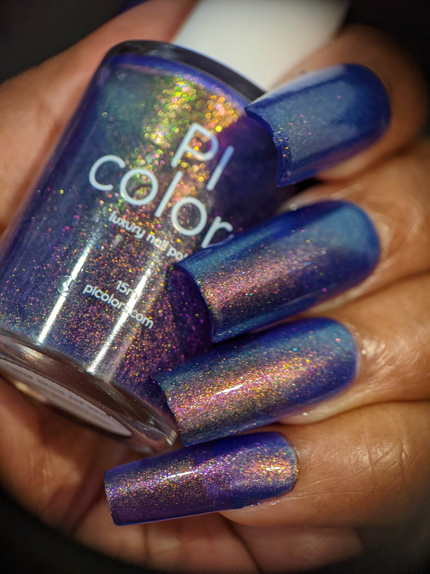 Close-up of a hand with long, blue nails showcasing a multicolor shimmer. The hand holds a bottle of PI Colors' handmade 90s Kind of World Purple Nail Polish, matching the nail color with sparkling iridescent flakes and hints of violet purple aurora pigment.