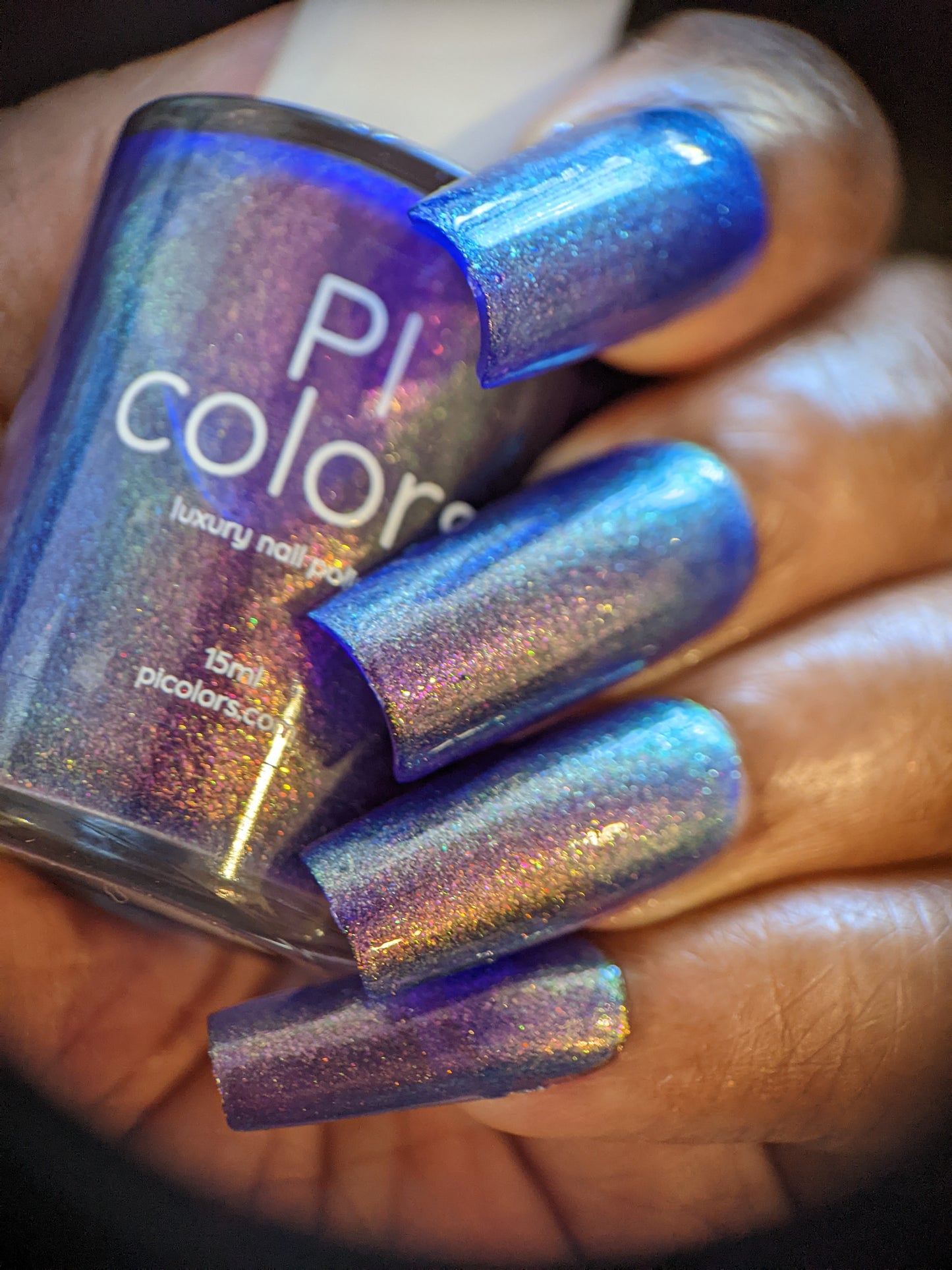 A close-up of a hand with long, oval-shaped nails painted in the shimmering hue of "90s Kind of World Purple Nail Polish," infused with aurora pigment and displaying holographic effects that reflect various colors under the light, while holding a bottle from PI Colors.