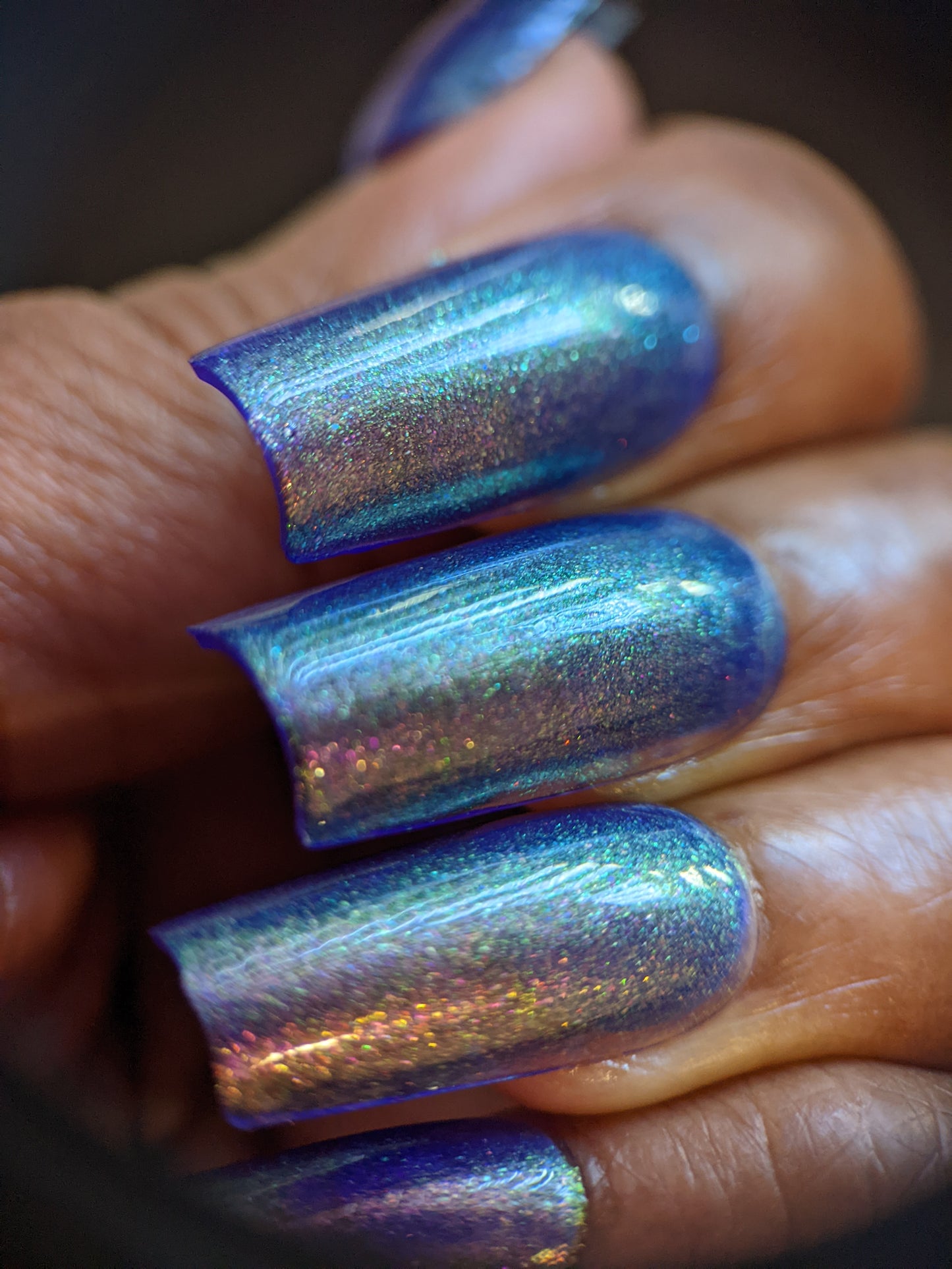 A close-up of a hand with long nails adorned in PI Colors' 90s Kind of World Purple Nail Polish. The shimmering blue and violet hues, enhanced by the aurora pigment, create a smooth, glossy finish that reflects light to produce a stunning rainbow effect on the surface. The skin tone is medium brown.
