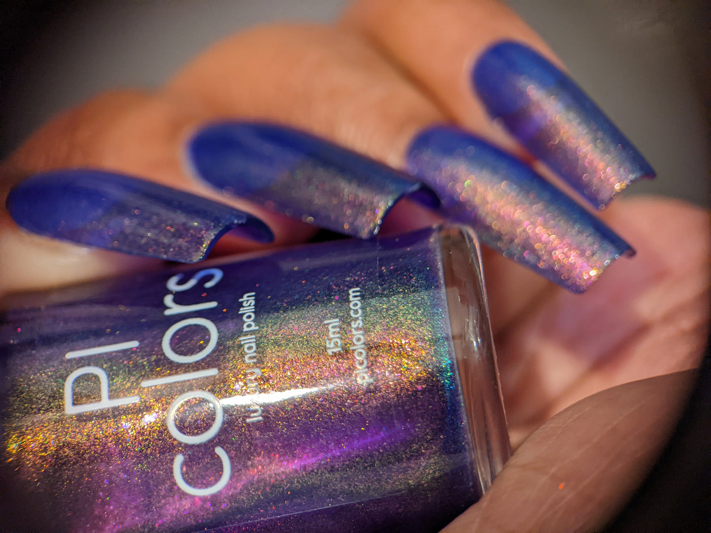 Close-up of a hand showcasing long nails painted with shimmering violet purple polish, featuring aurora pigment. The handmade nail polish bottle is held in the hand, revealing a matching color labeled "90s Kind of World Purple Nail Polish" by PI Colors.