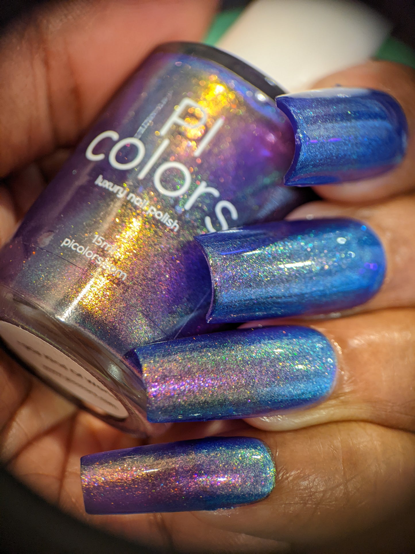 A hand showcases long nails painted with a shimmering, multicolored holographic purple infused with aurora pigment. The fingers hold a matching bottle of handmade nail polish labeled "90s Kind of World Purple Nail Polish" by PI Colors, with iridescent hues sparkling in the light.