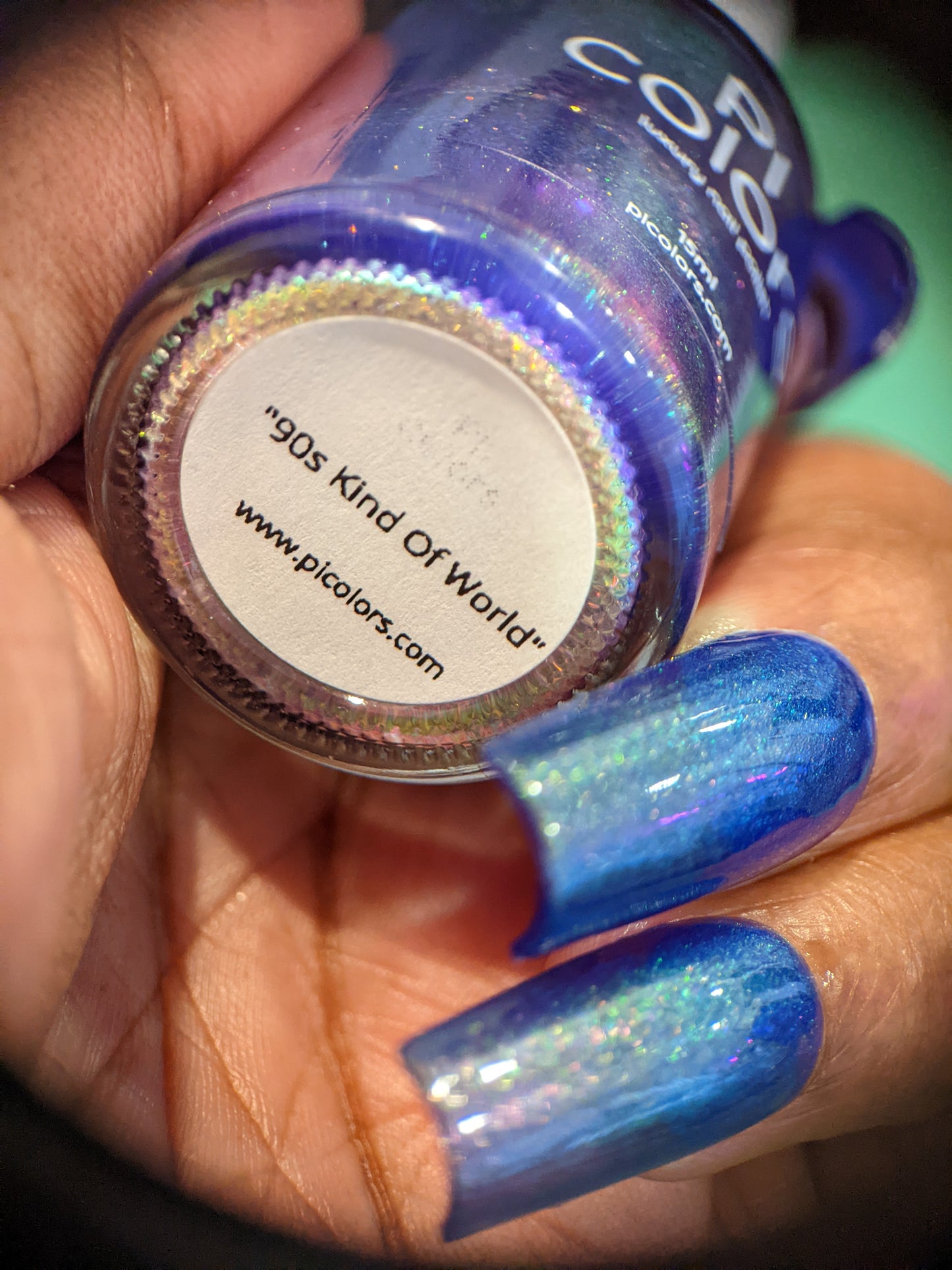 Close-up of a hand holding a purple nail polish bottle labeled "90s Kind of World" by PI Colors. The person's nails, painted with the same shimmery aurora pigment, reflect light and display a smooth, glossy finish.