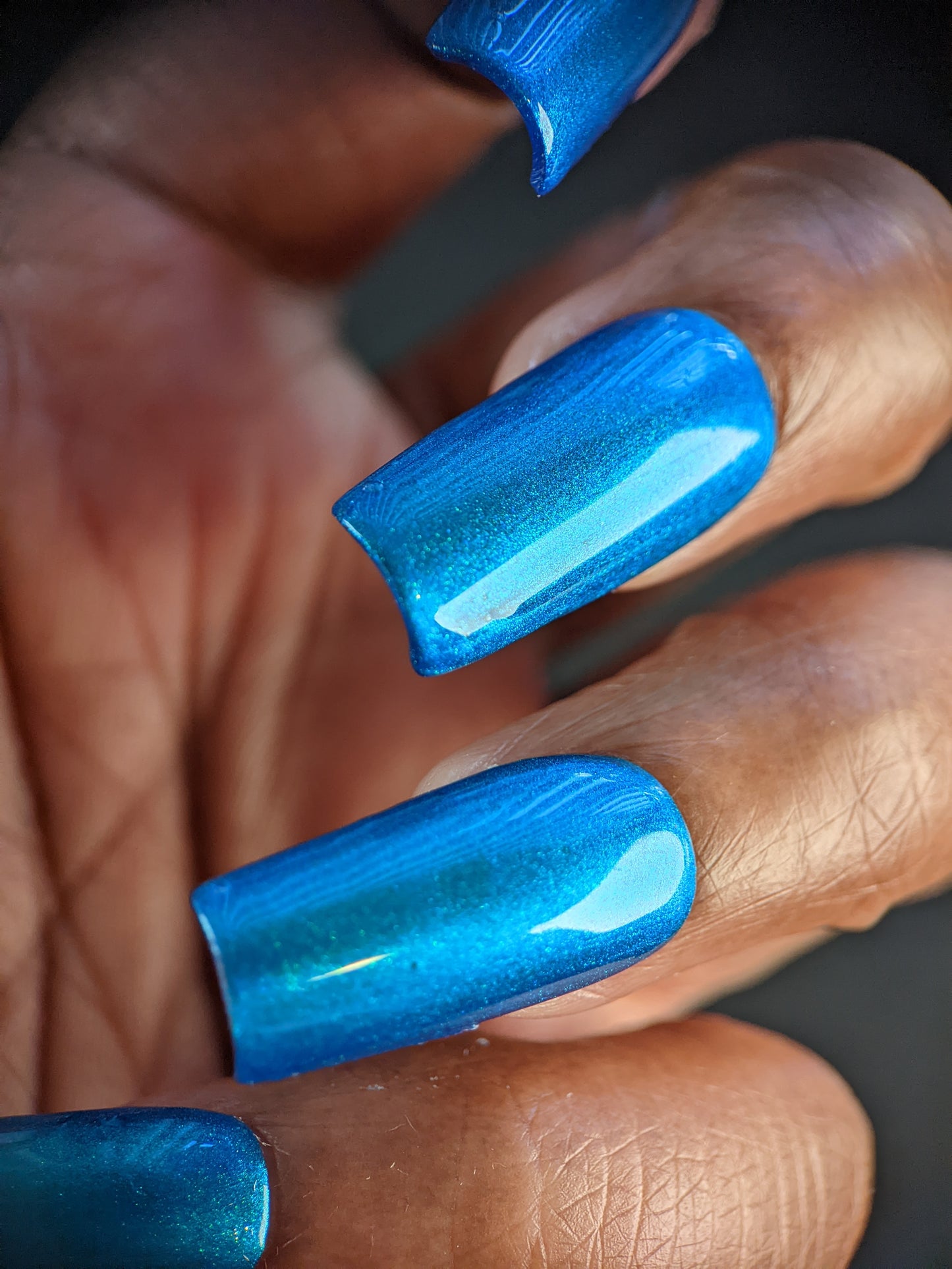 Close-up of a hand with smooth, glossy nails adorned in PI Colors' Prismatic Blue. The vivid color reflects light and boasts a subtle iridescent sheen, creating a striking visual effect against the skin tone—truly an indie masterpiece.