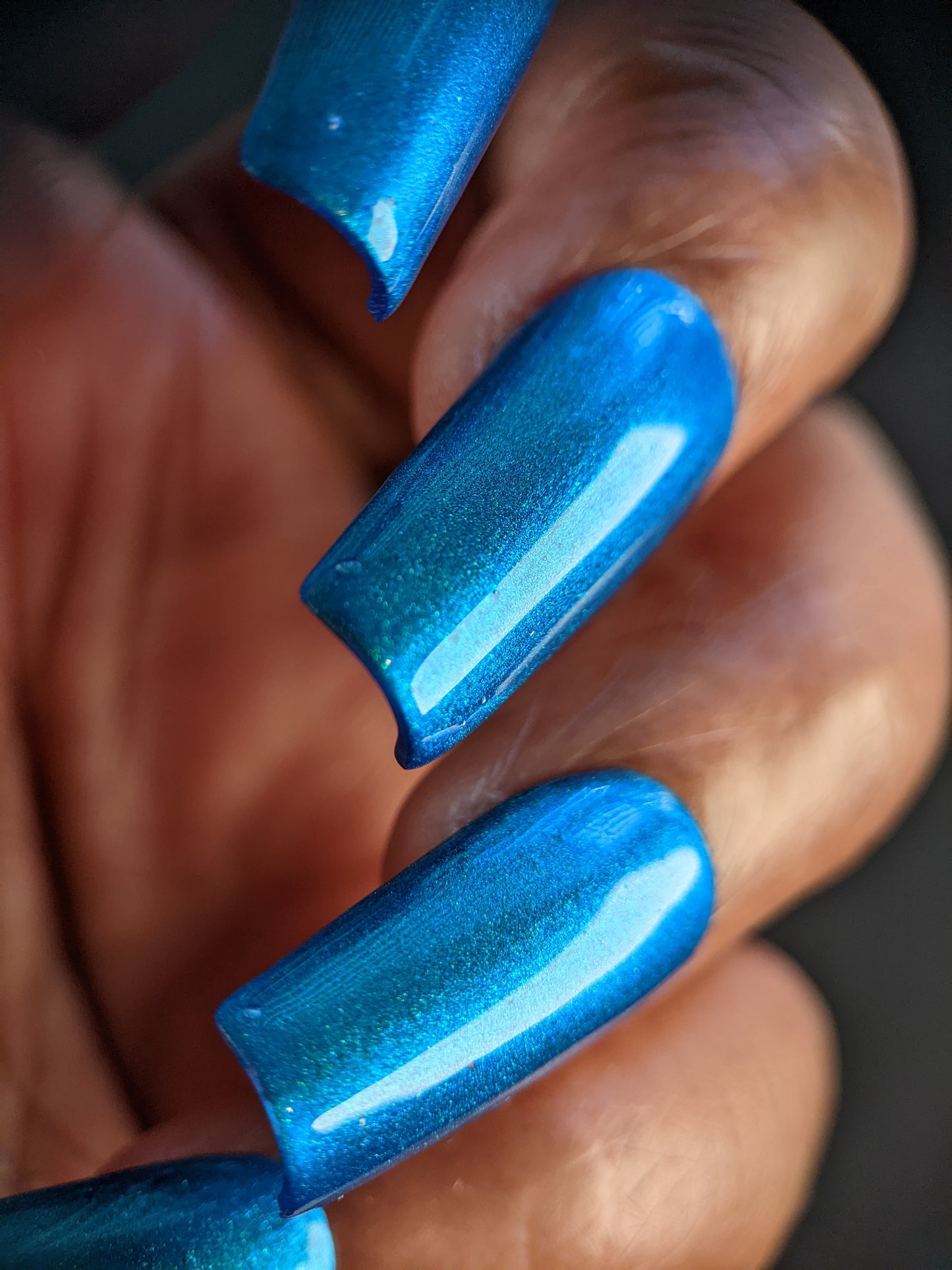 A close-up image showcases a hand decked out with long, shiny Prismatic Blue acrylic nails from PI Colors. The glossy finish reflects light brilliantly, and the vibrant color truly stands out against the dark, blurred background, exuding an indie vibe.