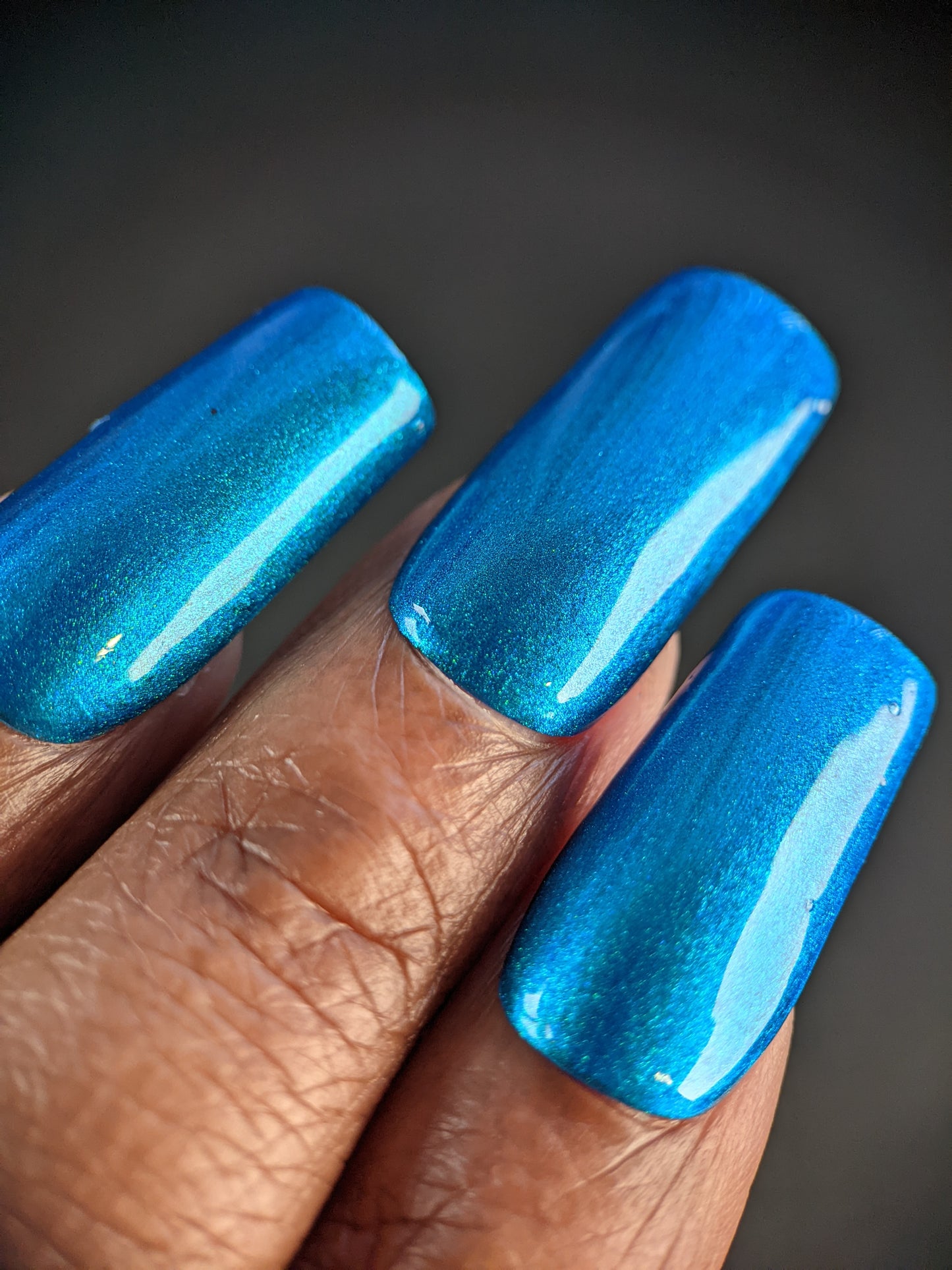 Close-up of a hand featuring long, glossy nails painted in Prismatic Blue. These 10-free nails have a smooth, reflective finish and glimmer beautifully under light. The blurred background accentuates the vibrant color of this nail polish crafted by PI Colors.