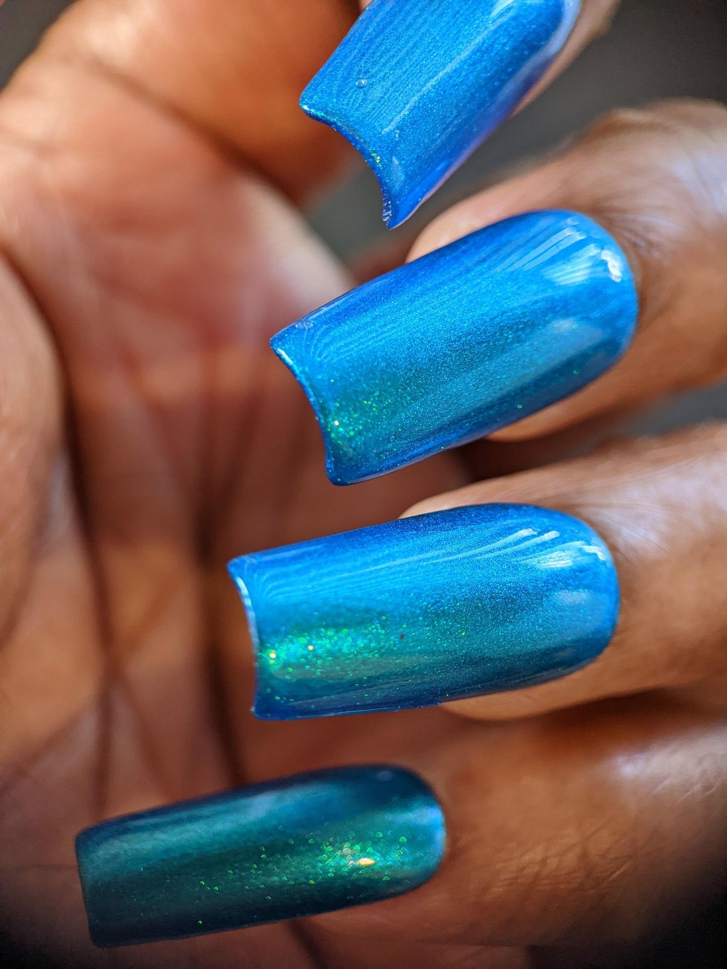 A close-up shot showcases a hand with long, square-shaped nails adorned in PI Colors' Prismatic Blue from their Luxury Nail Lacquers collection. The pearlescent finish captures and reflects light to reveal delicate green and teal undertones. On medium-dark skin, the glossy, polished nails exude elegance and style.