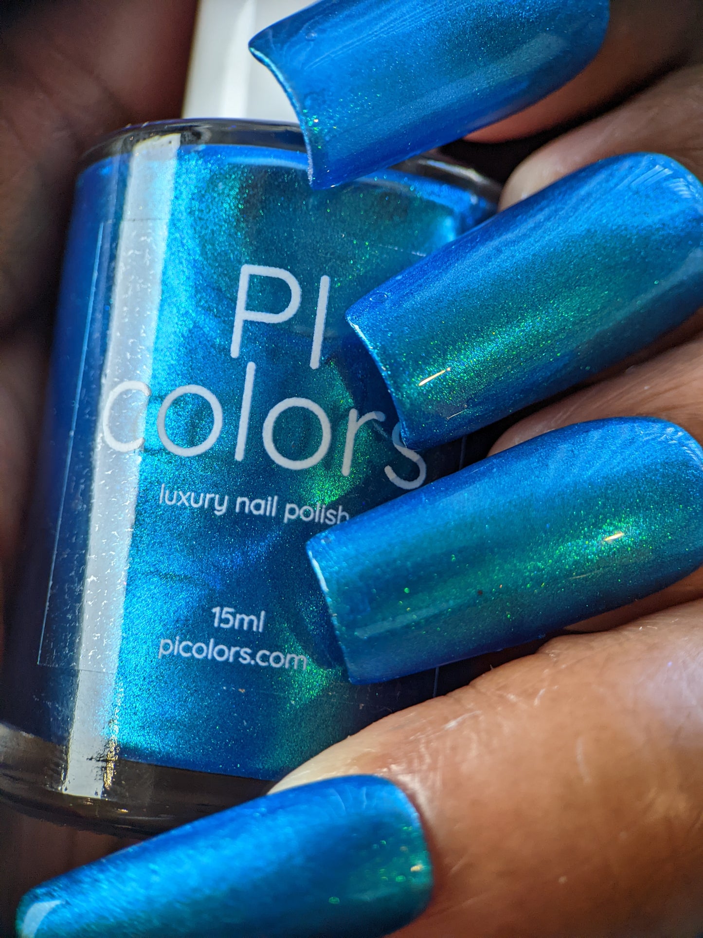 A close-up shot shows a hand with long nails painted in Prismatic Blue, gripping a bottle labeled "PI Colors Prismatic Blue Nail Polish, 15ml." This 10-free formula features a metallic and glossy finish, highlighting its place in the Indie Collective's stunning range.