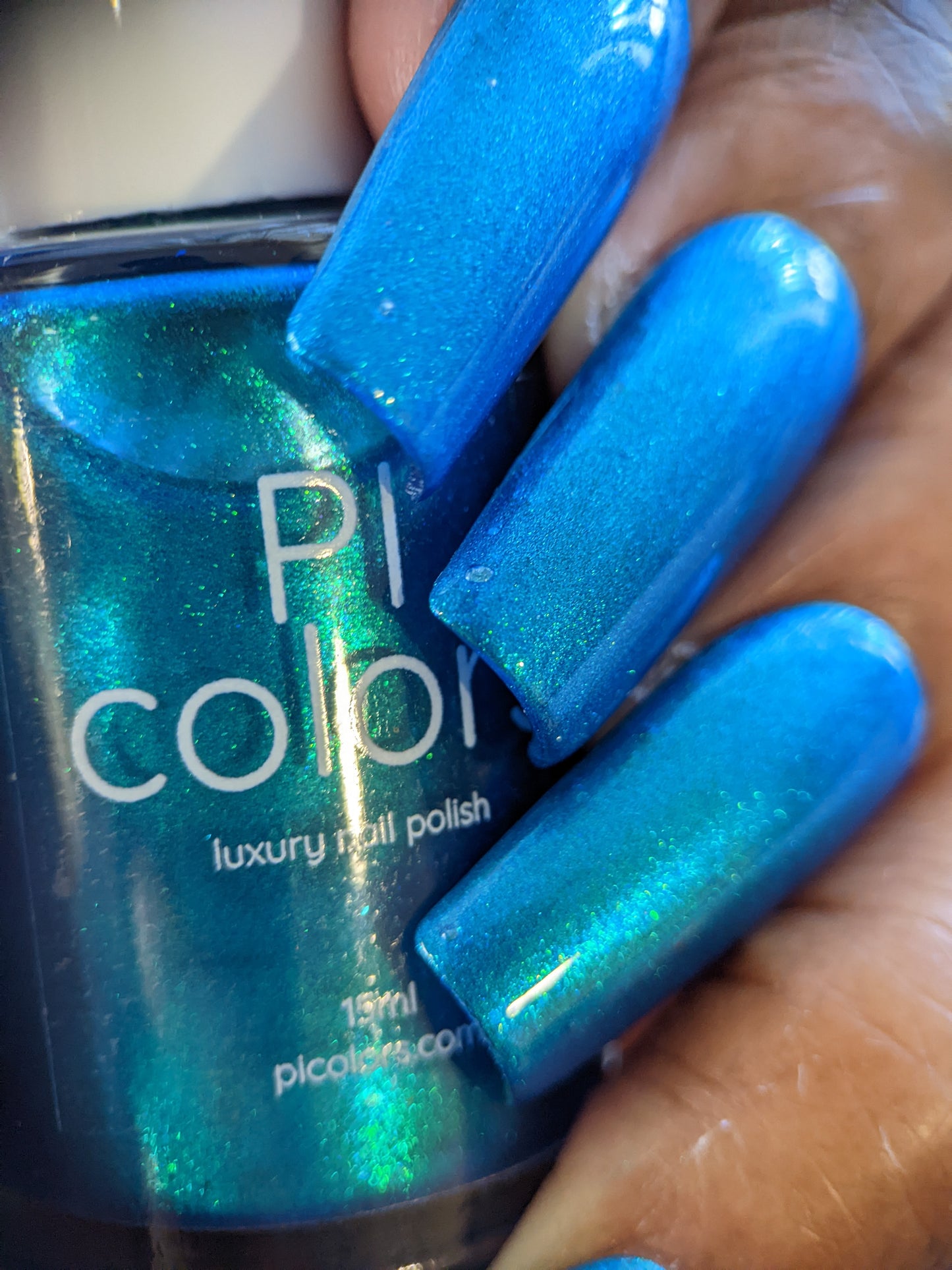 A close-up of a hand featuring long nails painted in Prismatic Blue nail polish, clutching a bottle labeled "PI Colors Luxury Nail Lacquers" in the same vibrant shade. The polish shines with a shimmering effect under light, embodying the essence of indie collective elegance.