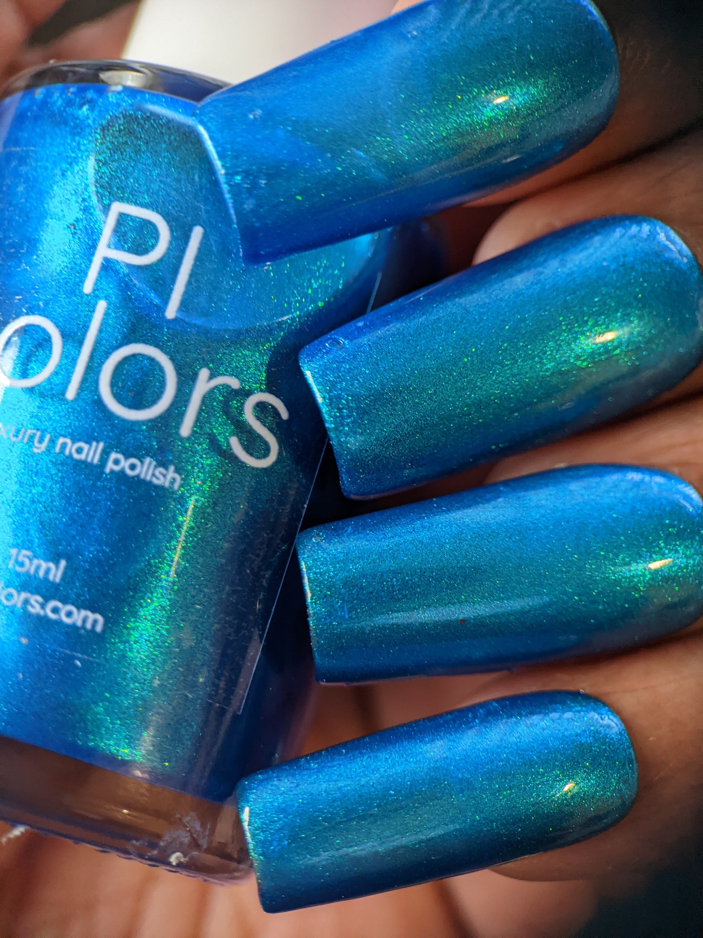 A hand adorned with long nails painted in Prismatic Blue nail polish holds a bottle of the same vibrant color. The polish bottle features the brand "PI Colors" and the text "Luxury Nail Lacquers" in white, embodying the essence of indie collective luxury.