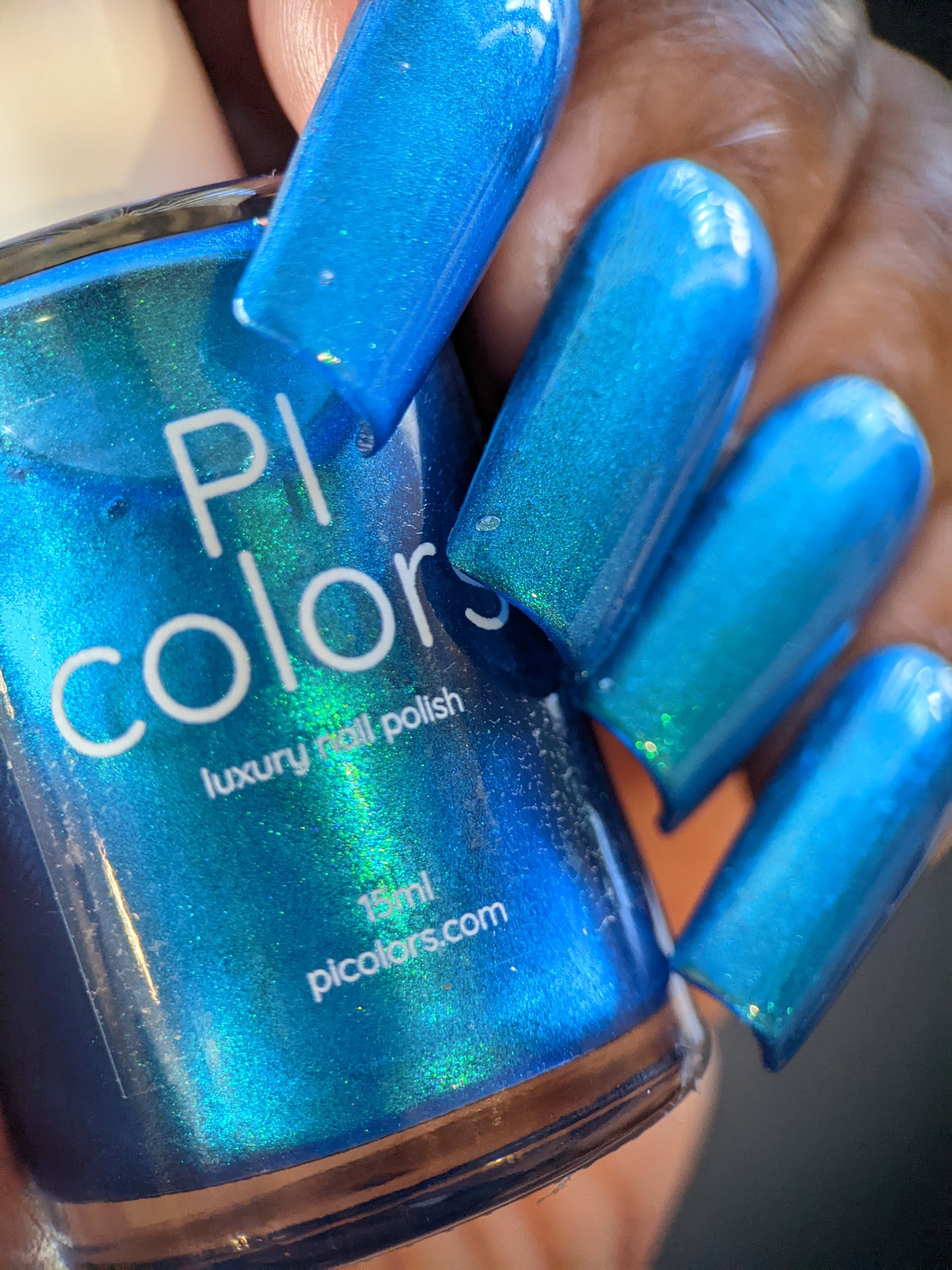 A close-up shot captures a hand with long, prismatic blue nails holding a bottle of PI Colors' Prismatic Blue nail polish. The polish perfectly complements the nail color with its shimmering effect, exemplifying the refined elegance typical of Luxury Nail Lacquers by Indie Collective.