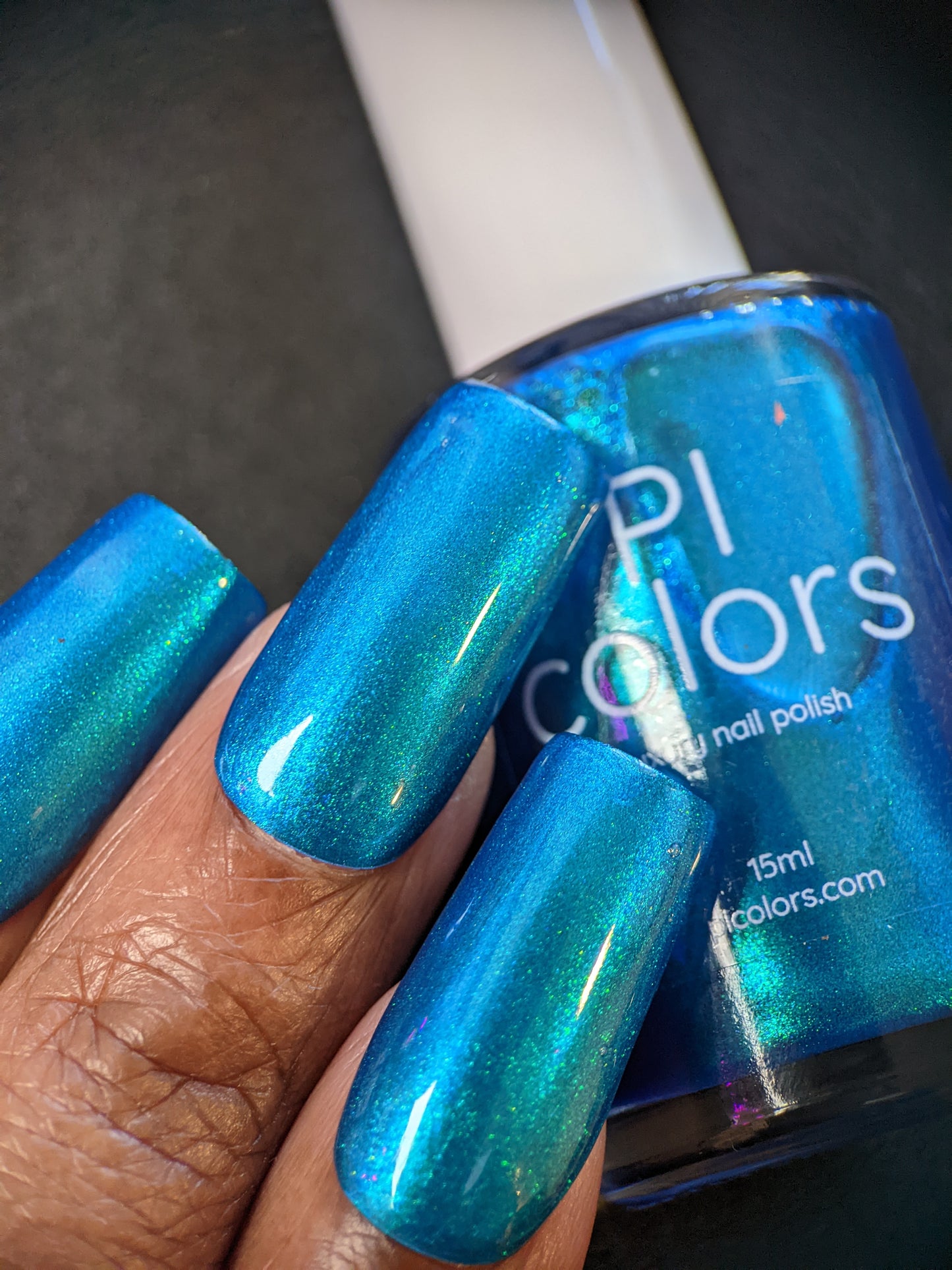 A close-up shows a hand showcasing long fingernails painted in Prismatic Blue, a shiny, metallic shade from PI Colors' Luxury Nail Lacquers collection. In the background is a bottle labeled "PI Colors," its vibrant hue beautifully contrasted against the dark backdrop.