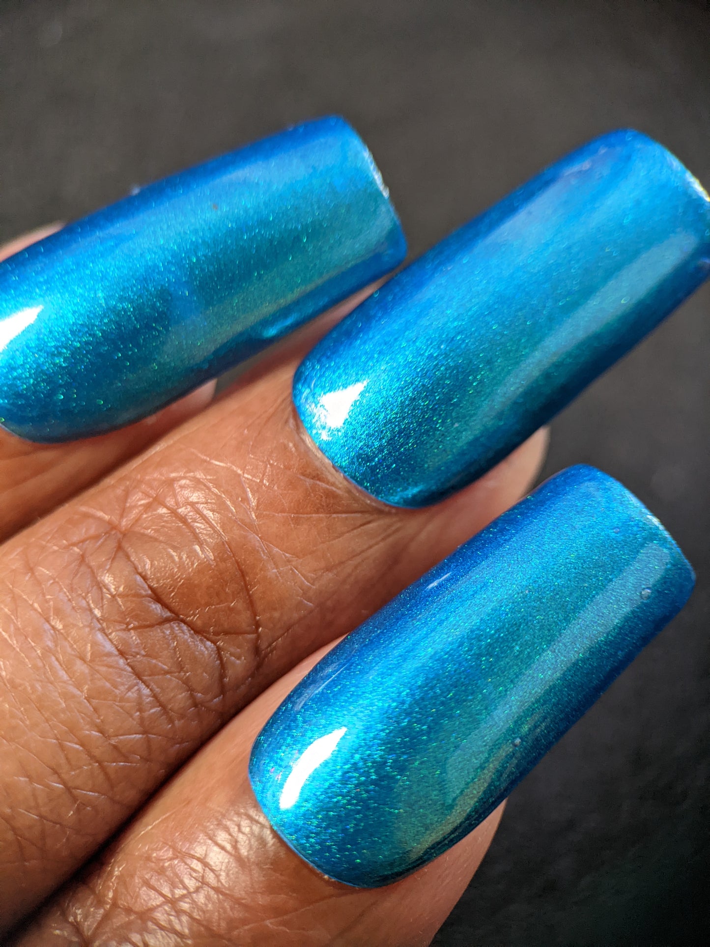 Close-up of a hand displaying long, glossy nails coated with Prismatic Blue by PI Colors. The shimmering finish of this 10-free polish beautifully complements the warm skin tone against a dark background.
