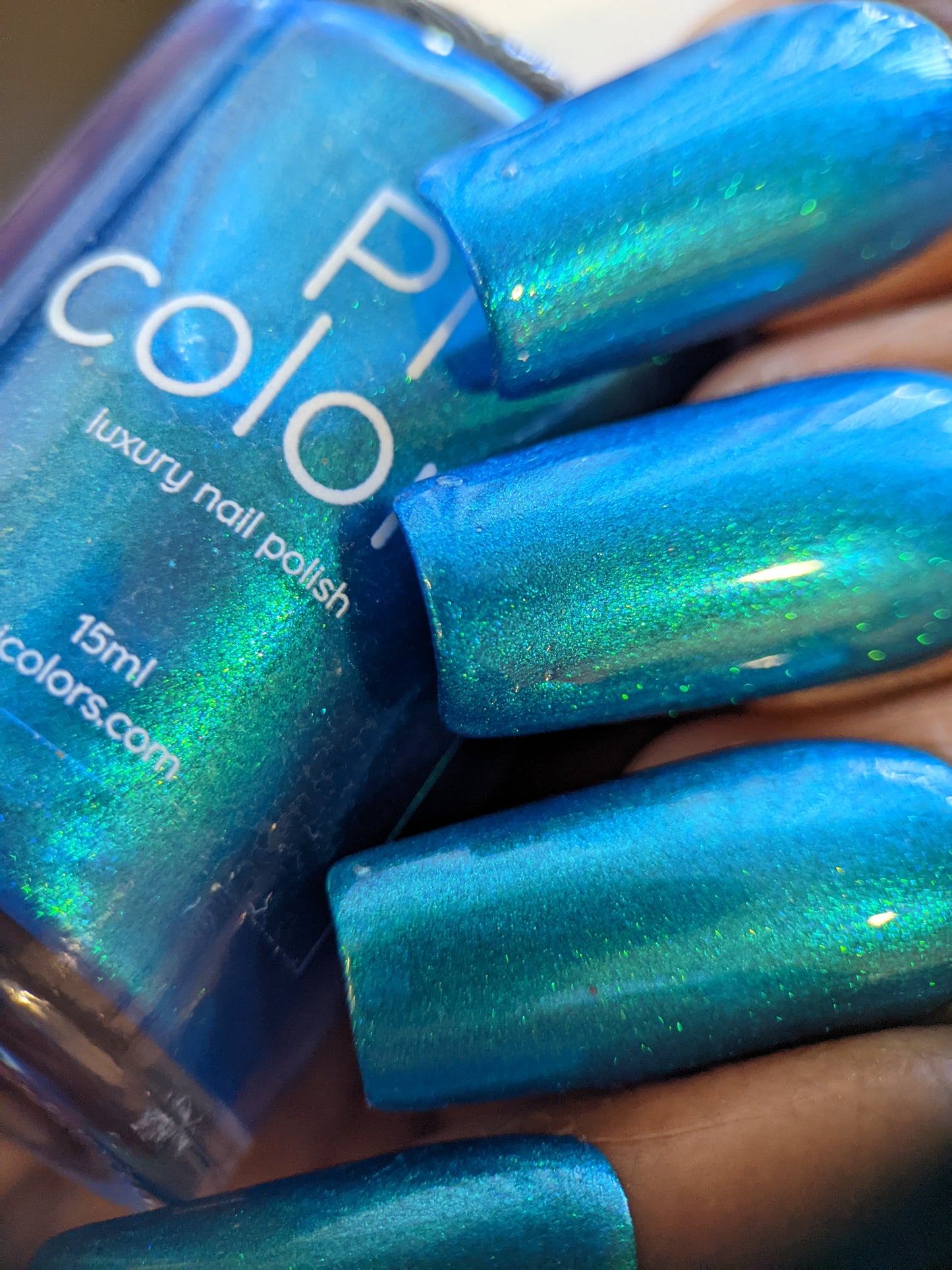 A close-up of four fingernails painted with the shimmering Prismatic Blue from PI Colors, holding a bottle of Luxury Nail Lacquers labeled "15ml, luxury nail polish." The polish boasts a glossy, iridescent finish.