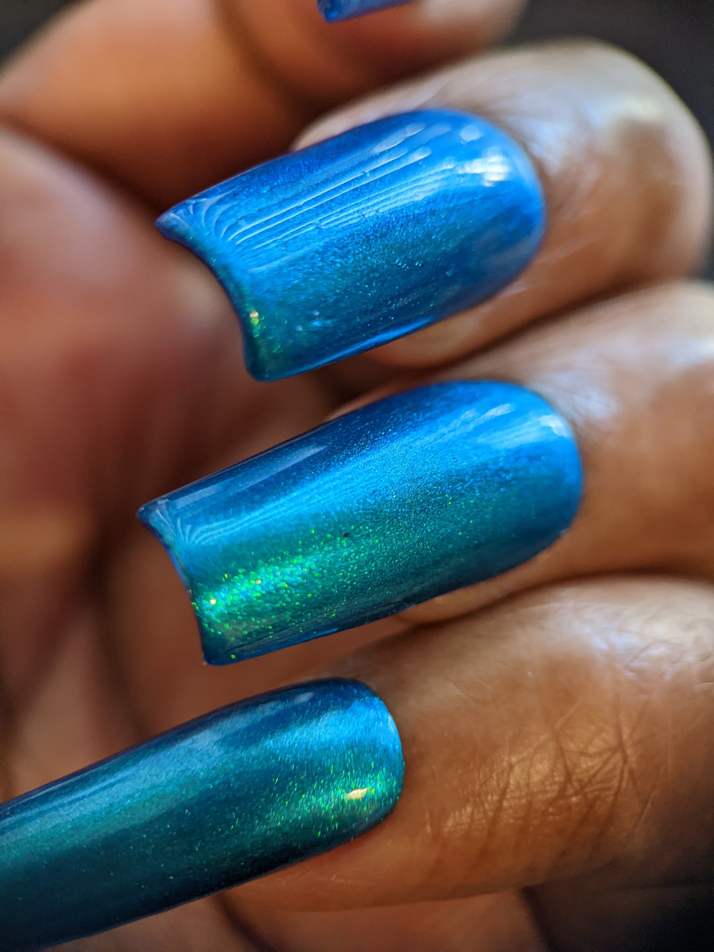 A close-up shows long, glossy nails painted with PI Colors' Prismatic Blue. This 10-free polish features a metallic sheen and subtle green iridescence, creating a striking contrast against the partially visible skin and embodying an Indie Collective style.
