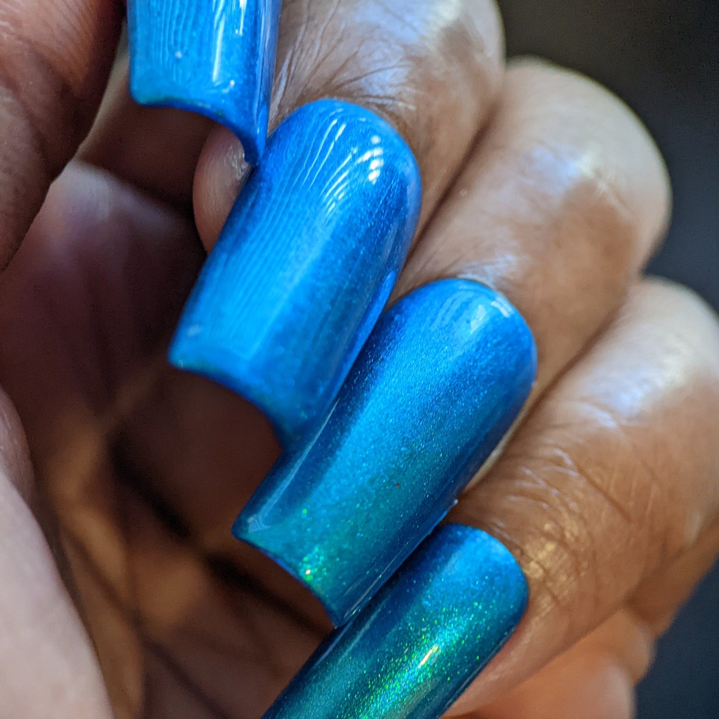 A close-up of a hand showcases the Prismatic Blue nail lacquer by PI Colors, featuring long, captivating blue nails with a glossy, reflective finish. This luxurious nail polish catches the light, revealing subtle shade variations against warm skin tones and a softly blurred background.
