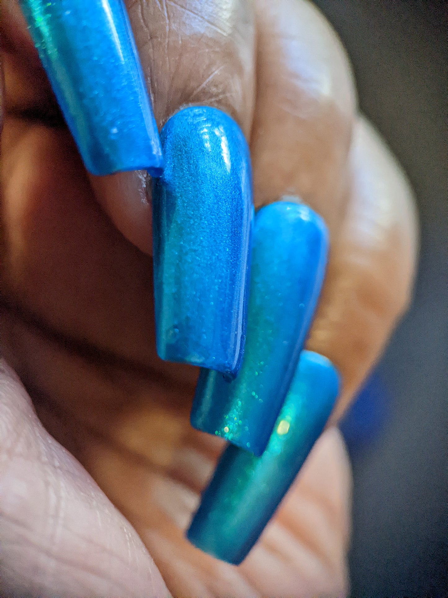 Close-up of a hand with long, glossy PI Colors nails in Prismatic Blue. This luxury nail lacquer features a metallic sheen that brilliantly reflects light. The blurred background draws attention to the vibrant Indie Collective hue.