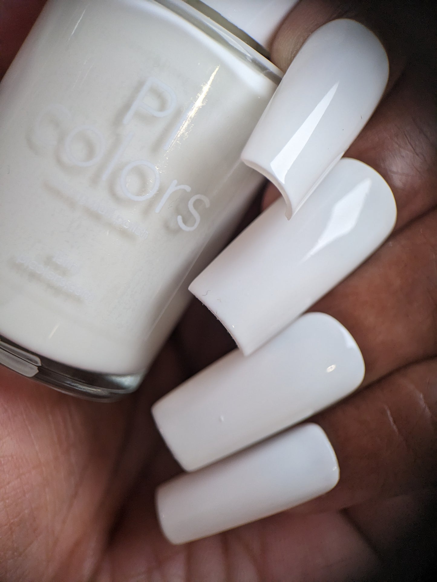 A close-up view shows a hand holding a bottle of PI Colors Bright White.223 Nail Polish. The nails boast a glossy crème finish in matching white, featuring an elegant square-shaped long manicure.