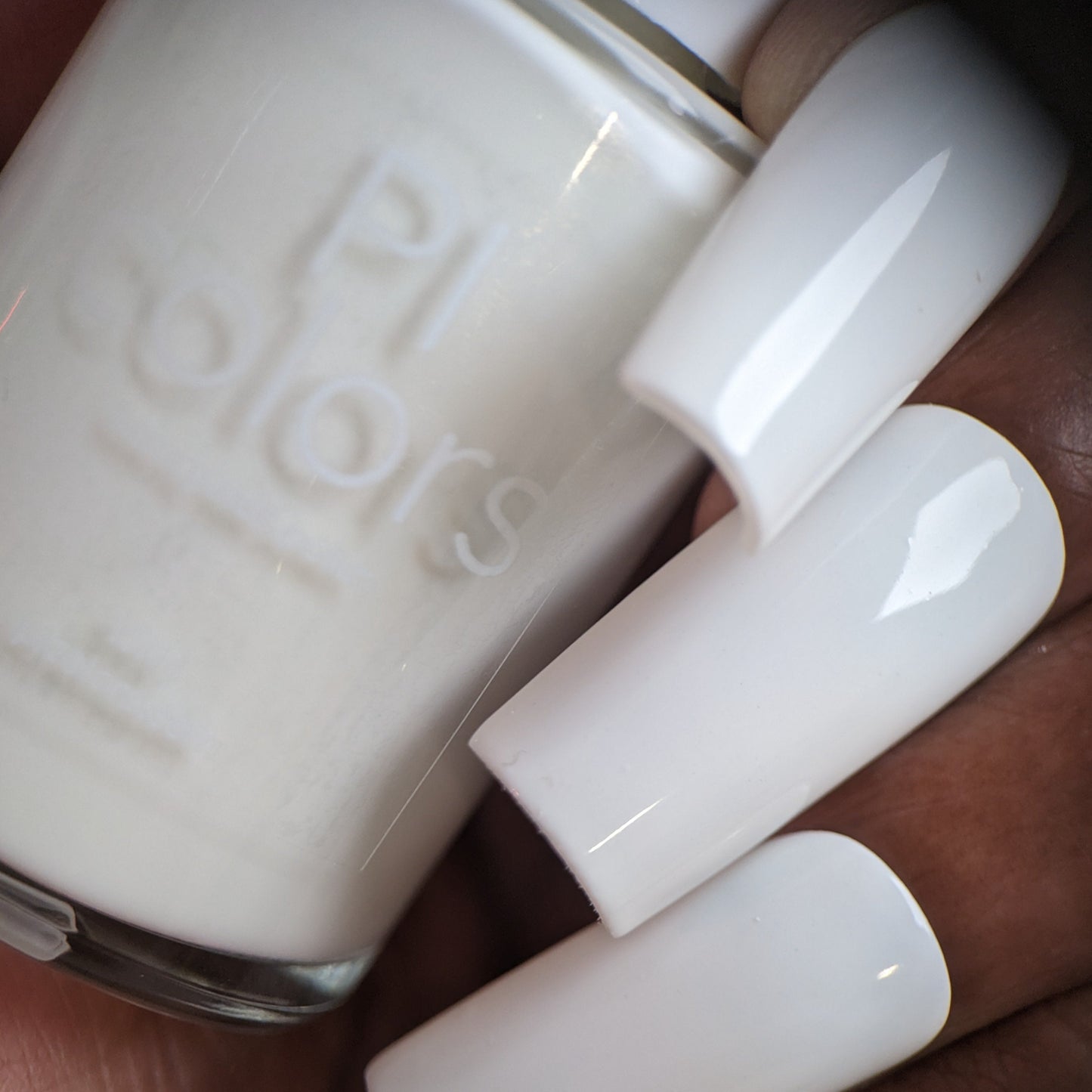 Close-up of a person holding a bottle of Bright White.223 White Nail Polish with long, polished nails that sport a glossy creme finish. The bottle reads "PI Colors," and its 10-free formula ensures the nails match perfectly.