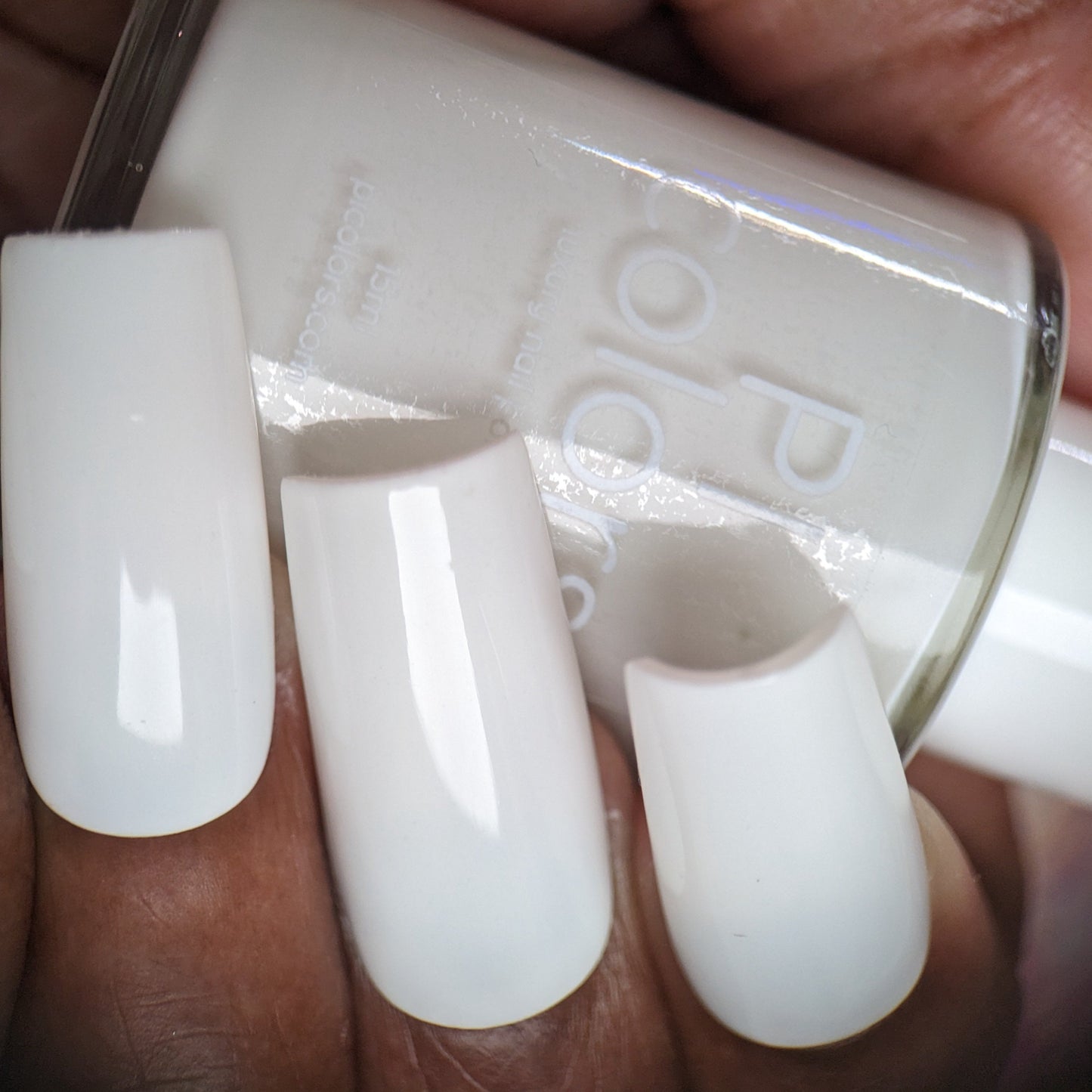 A close-up captures a hand holding a bottle of PI Colors Bright White.223 White Nail Polish. The nails, painted in the same glossy crème finish, boast a smooth and shiny allure.