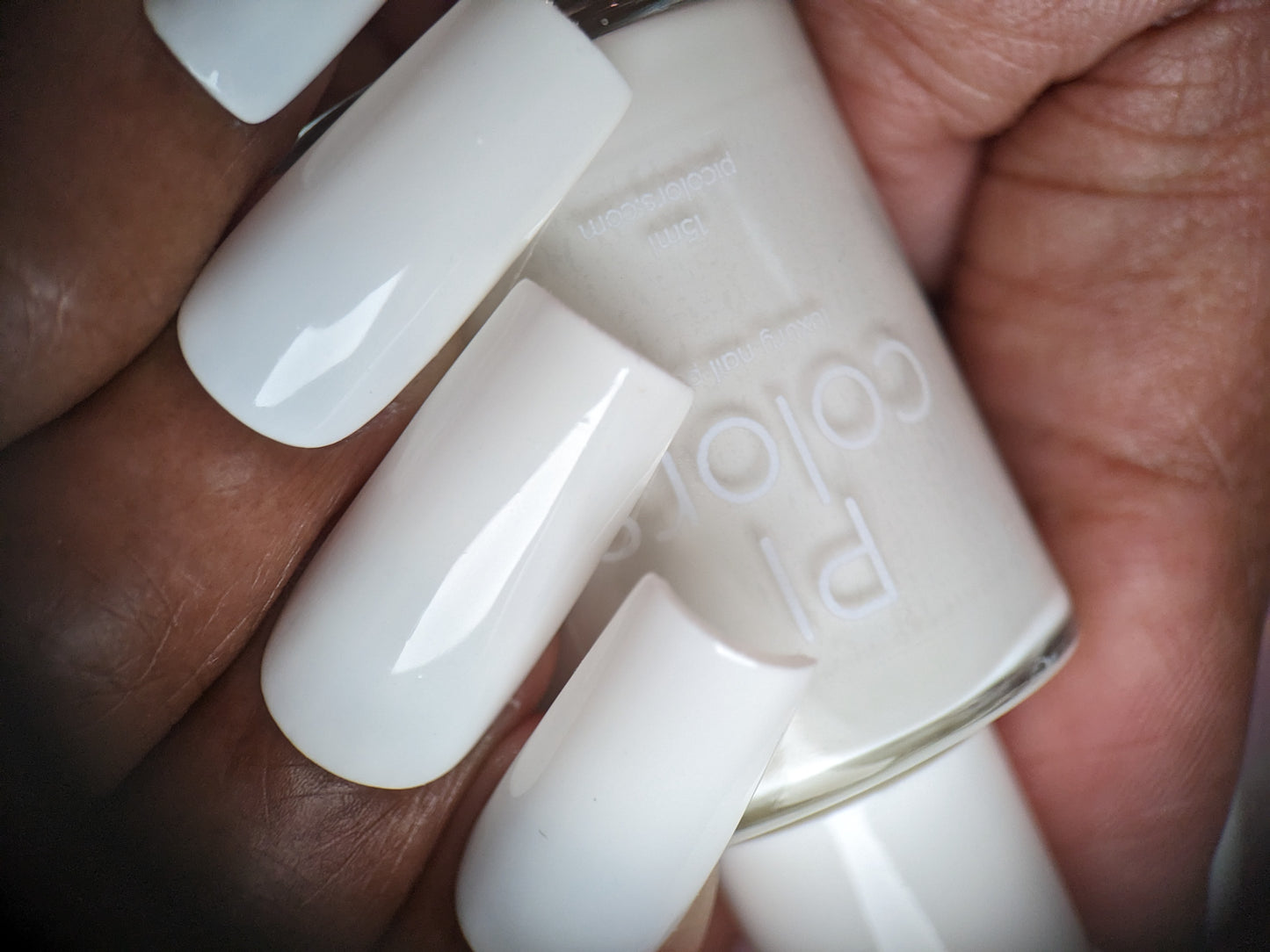 A close-up shot captures a hand with long, manicured nails painted in a glossy white hue, perfectly showcasing their elegance as they hold a bottle of PI Colors' Bright White.223 White Nail Polish, which matches the exquisite glossy creme finish on the nails.