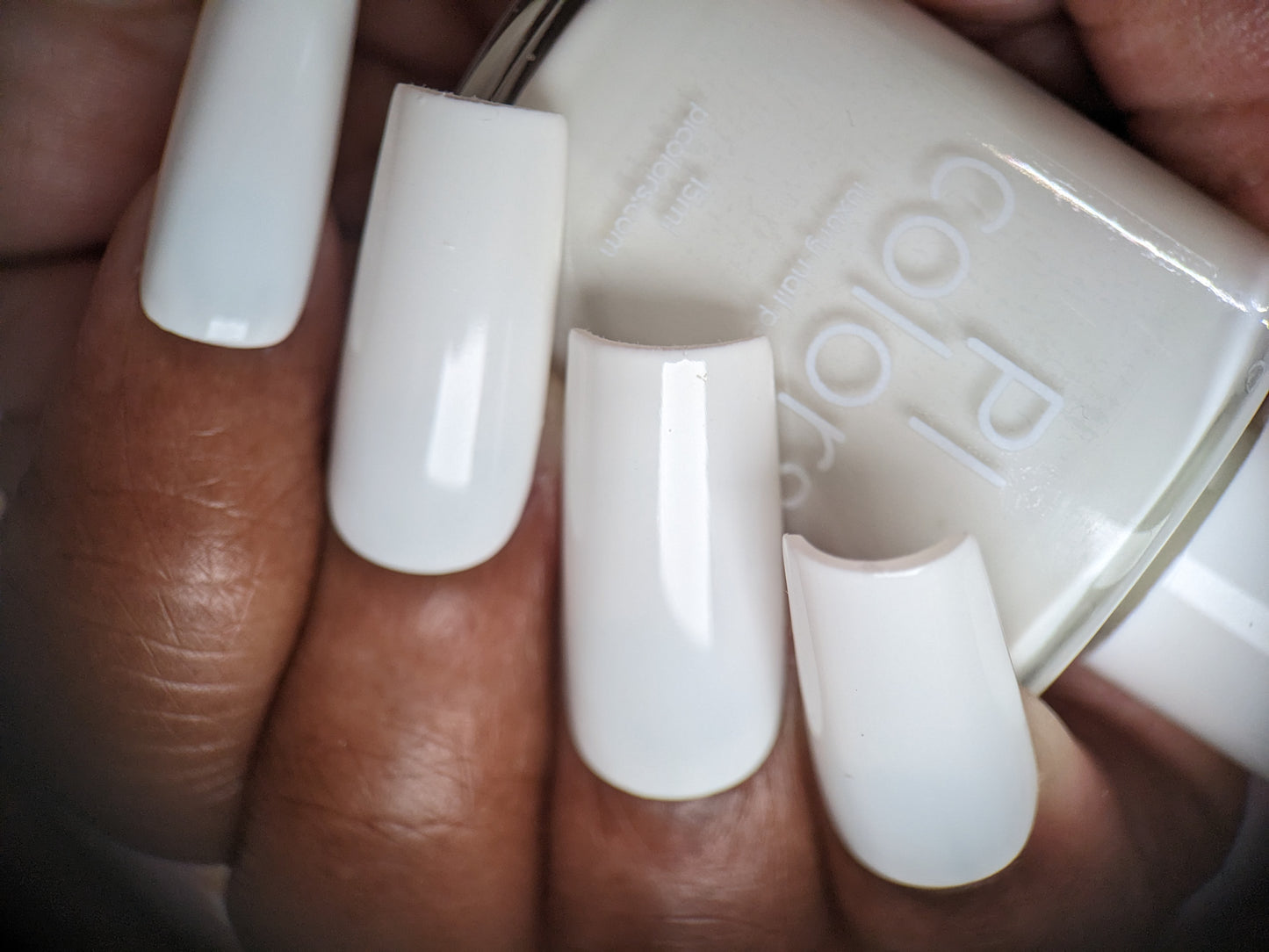 A hand with five long, polished nails holds a PI Colors-labeled bottle of Bright White.223 white nail polish. The nails are painted in a glossy creme finish, perfectly matching the bottle.